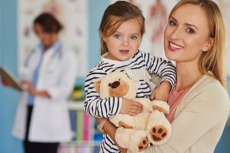 Mother and young child with a teddy bear in a bright clinic; a doctor studies a chart in the background, smiling at the camera.